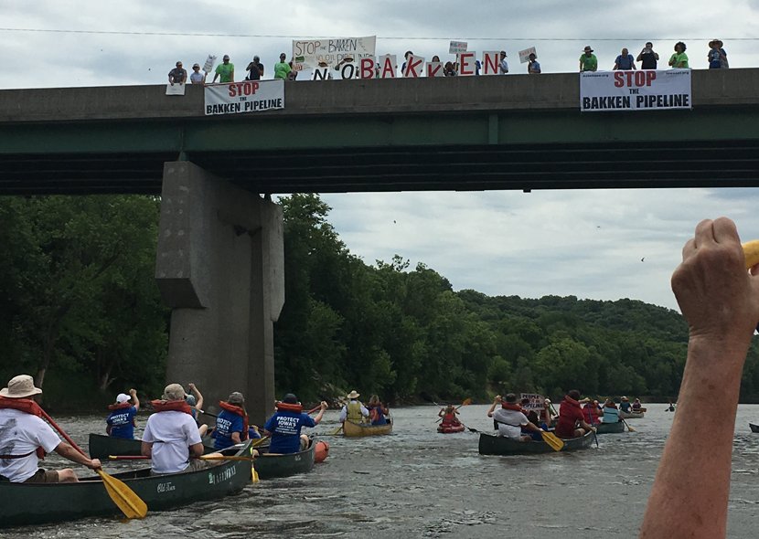 Several canoes filled with protestors sail down a river. Above them, on a bridge, more protestors hold banners that read "Stop the Bakken Pipeline."